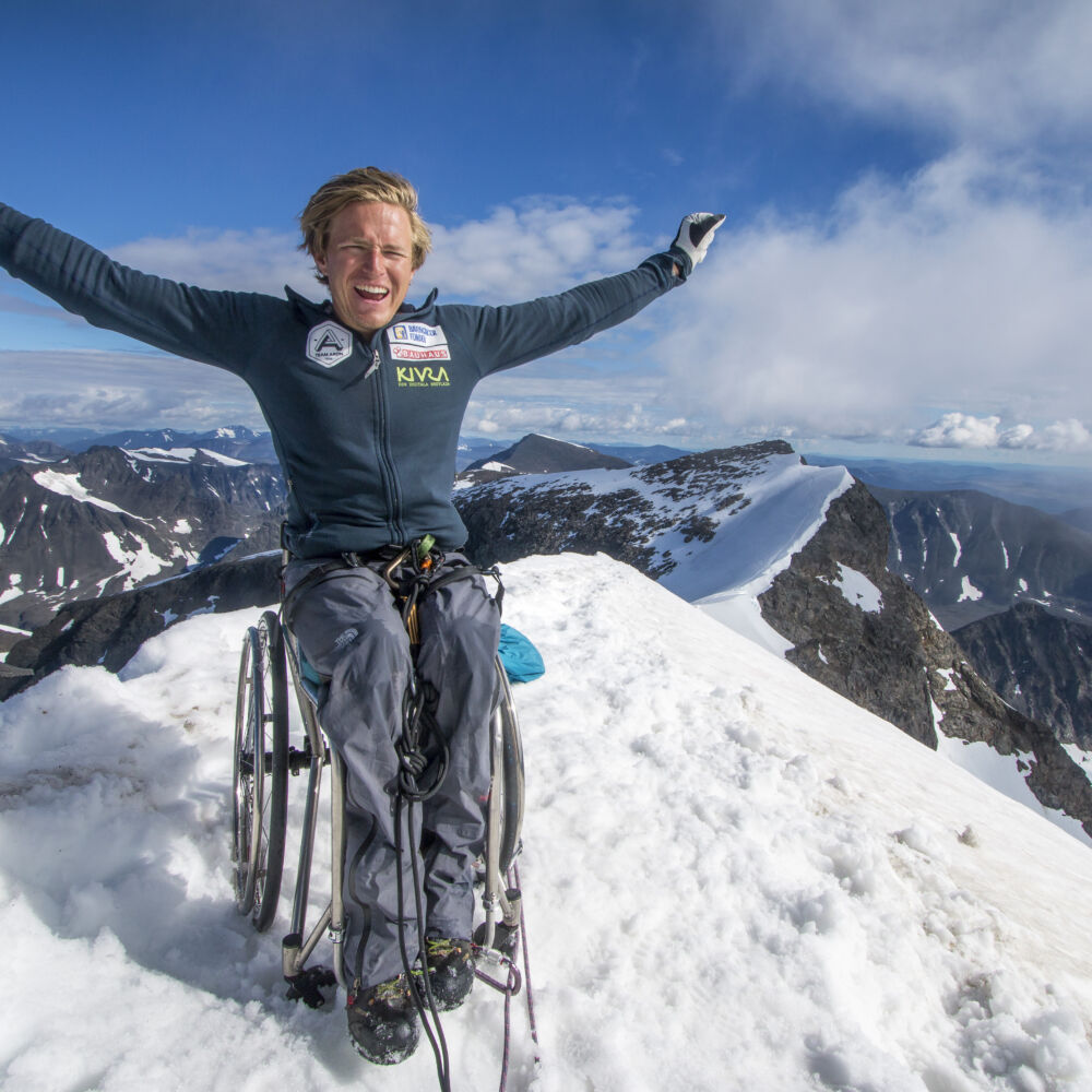 en man på rullstol stor med händerna upp på bergstopp täckt av snö