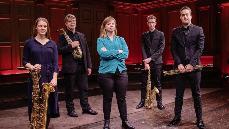 Fem personer står framför scenen. Fyra är musiker i svarta kläder med blåsinstrument. En kvinna i blå kavaj utan instrument.