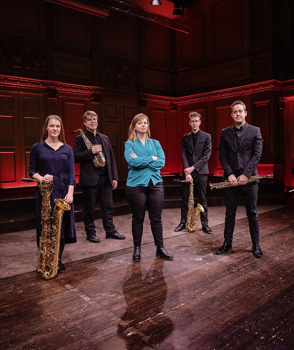 Fem personer står framför scenen. Fyra är musiker i svarta kläder med blåsinstrument. En kvinna i blå kavaj utan instrument.