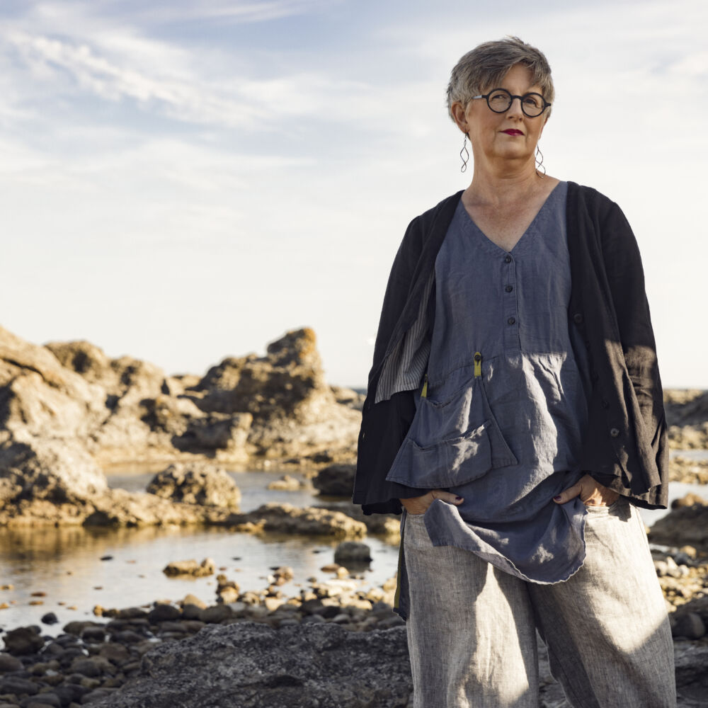 en kvinna i glasögon, och blå kläder står på stranden med stenar bakom