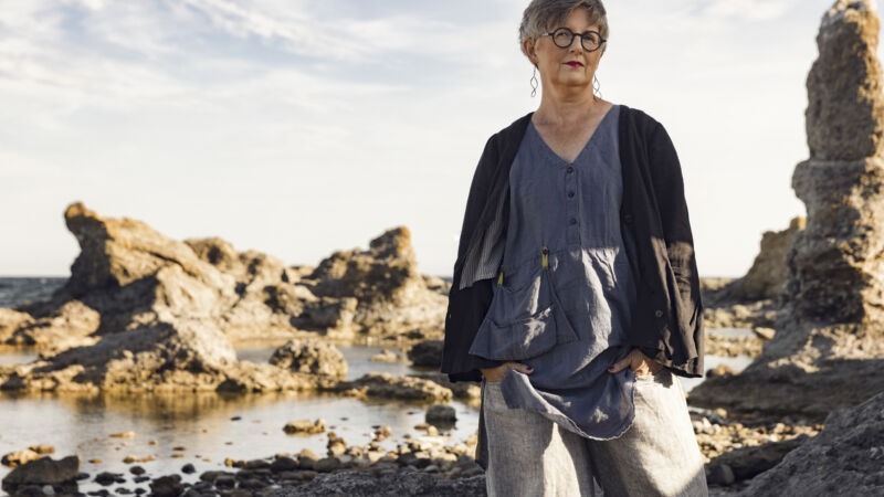 en kvinna i glasögon, och blå kläder står på stranden med stenar bakom