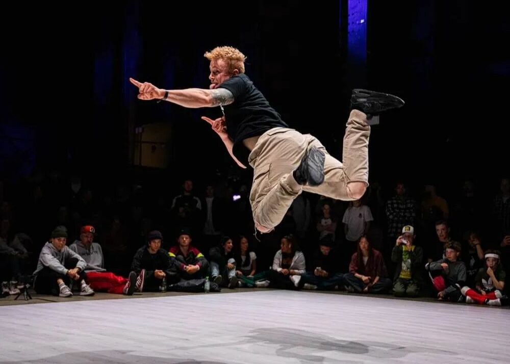 en blond man i en svart tröja och beige byxor dansar breakdance