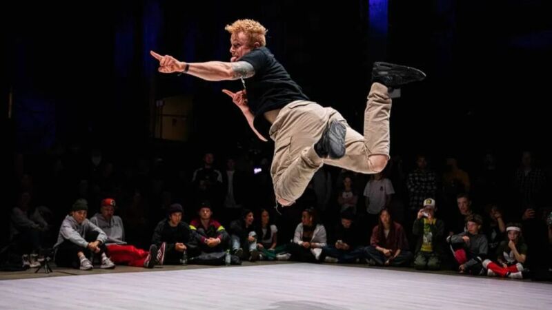 en blond man i en svart tröja och beige byxor dansar breakdance