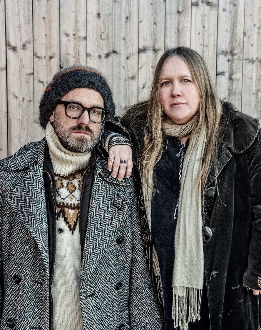 en man i en mössa och glasögon står med en blond kvinna i en svart kapp, bakom finn trävägg