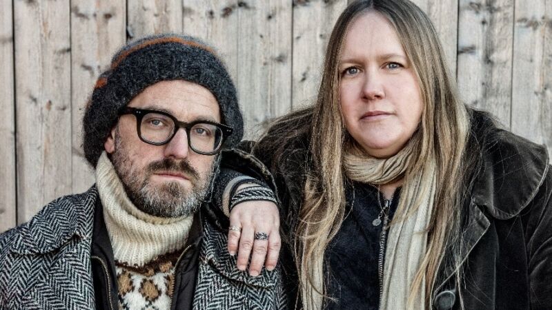 en man i en mössa och glasögon står med en blond kvinna i en svart kapp, bakom finn trävägg