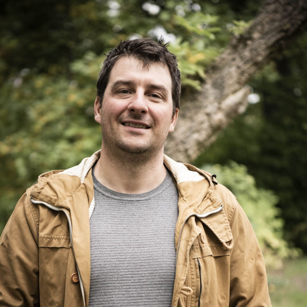 en man i medelålder, brun hår, en grå T-shir it, en gul jacka, i handen håller en bok Gäddbibbeln med en fiske på omslaget