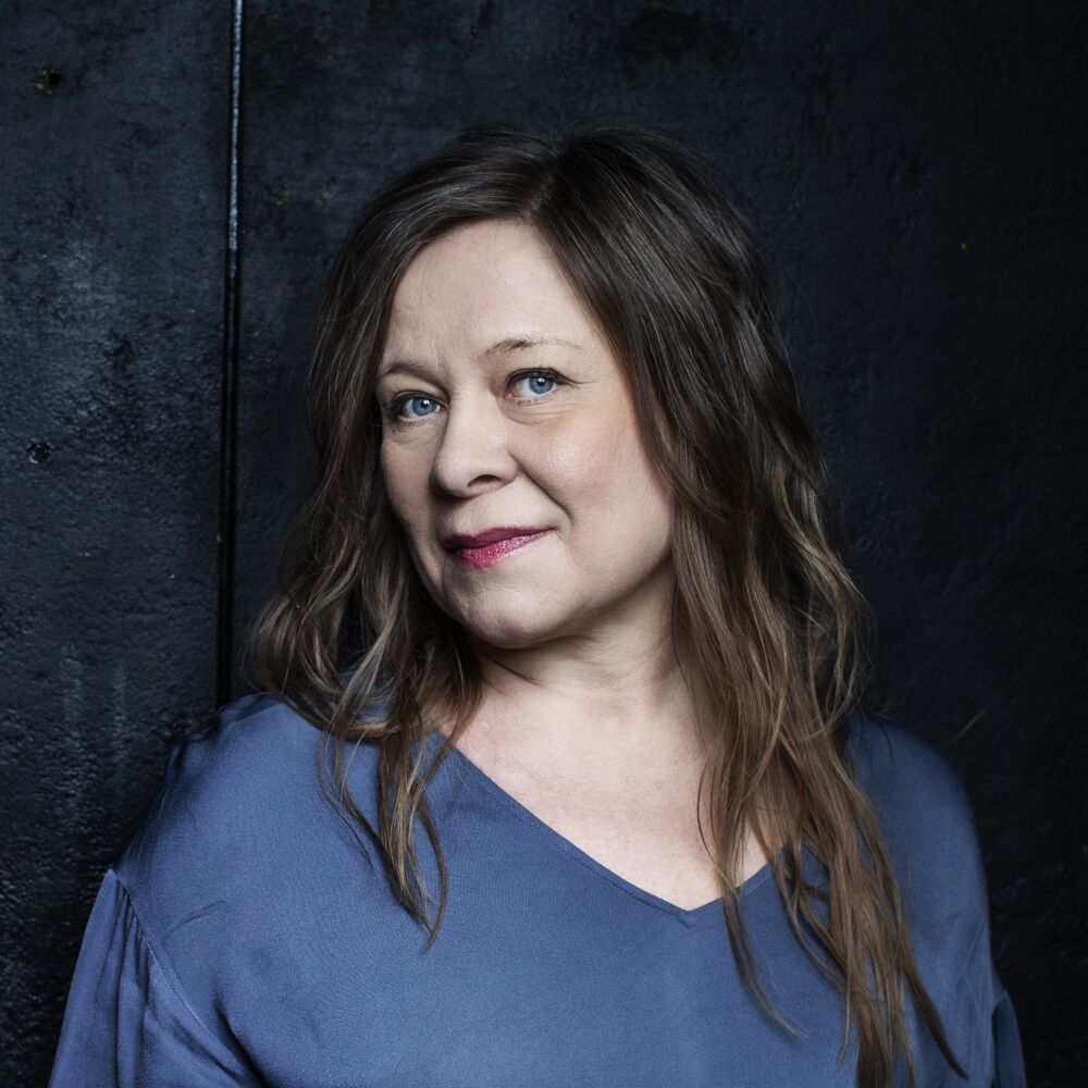 em kvinna med långt ljus hår och i en blå tröja mot svart bakgrund