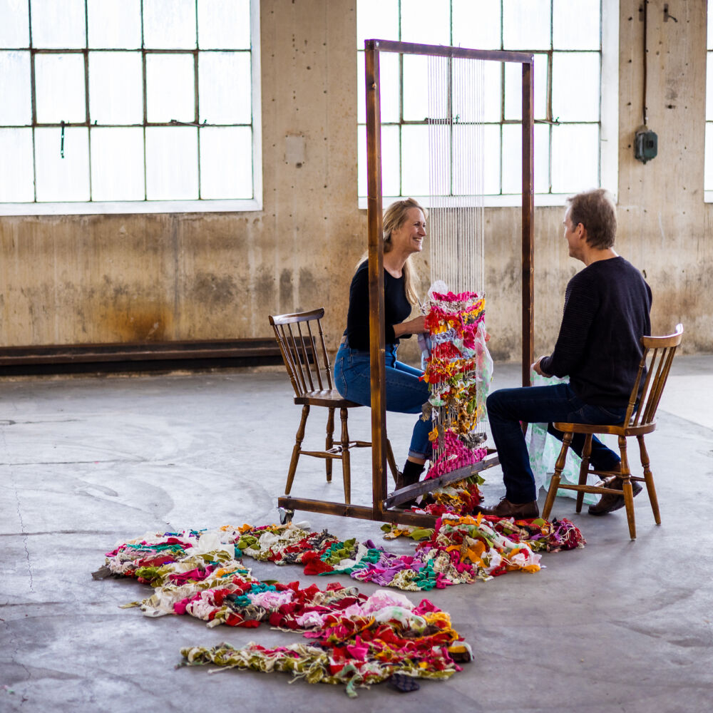 Två personer sitter i ett rum med en väv mellan dem. Vävt tyg ligger på golvet.