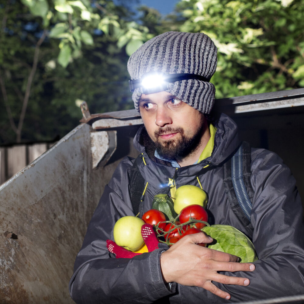 en man i en grå mössa och pannlampa håller grönsaker medan han kliver ur ut container