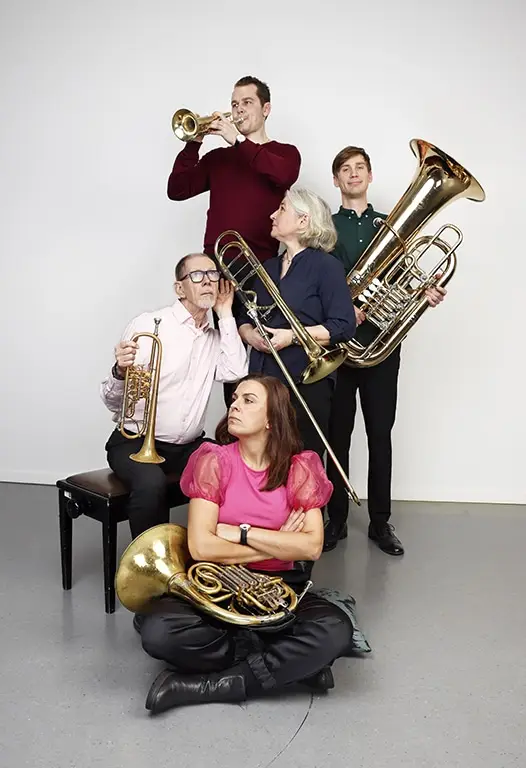 Bilden visar fem personer som spelar blåsinstrument som trumpet, trombon, tuba och valthorn. De är arrangerade i en grupp, där några står och andra sitter. Den vita bakgrunden gör att fokus ligger helt på musikerna och deras instrument.