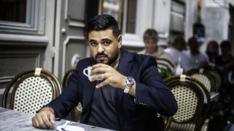 en man med mörk hår och i en blå kavaj sitter utomhus och dricker kaffe