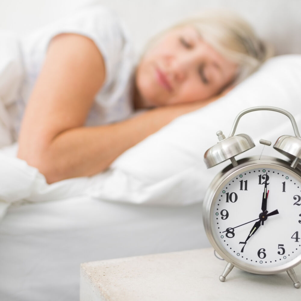 en kvinna med grå hår sover i sängen, på sängbordet ligger en klocka