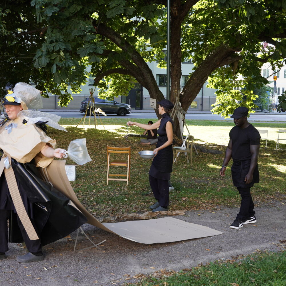 En person iklädd plast och kartong går på en grusväg. Efter sig har hon tre svartklädda personer som följer efter. Det är soligt väder.
