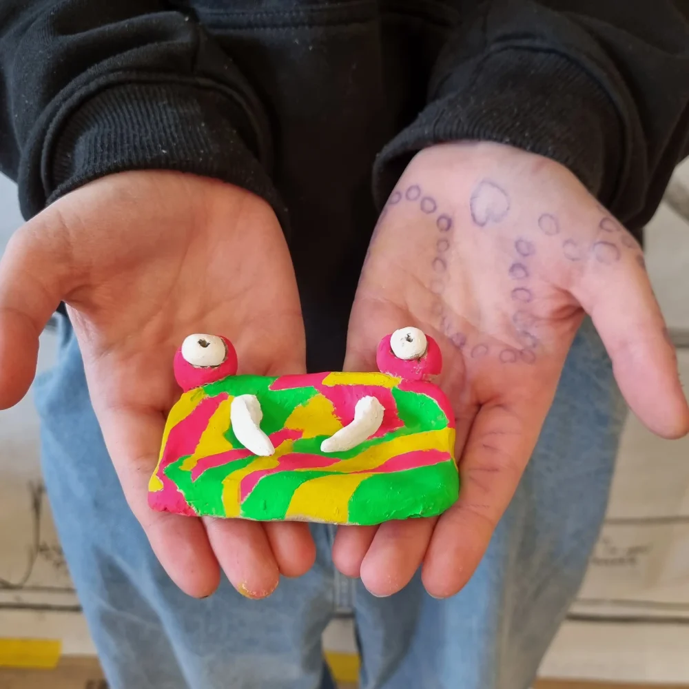 Två öppna händer som håller fram en färgglad figur som liknar en groda
