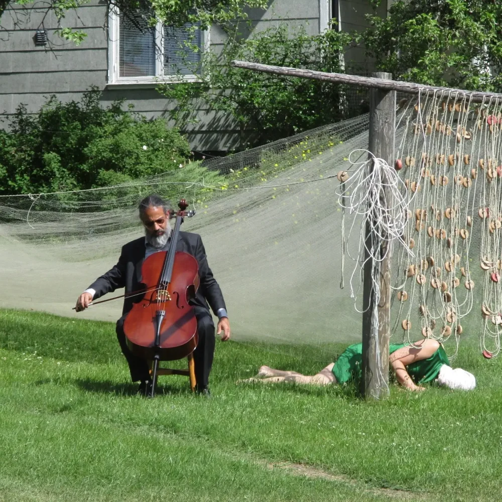 en person spelar cello på gräsmattan. ett fiskenät hänger i bakgrunden. en person ligger på gräsmattan.