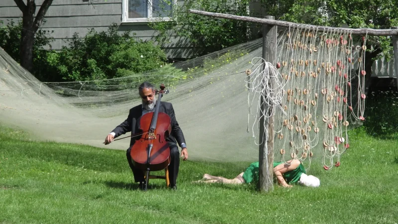 en person spelar cello på gräsmattan. ett fiskenät hänger i bakgrunden. en person ligger på gräsmattan.