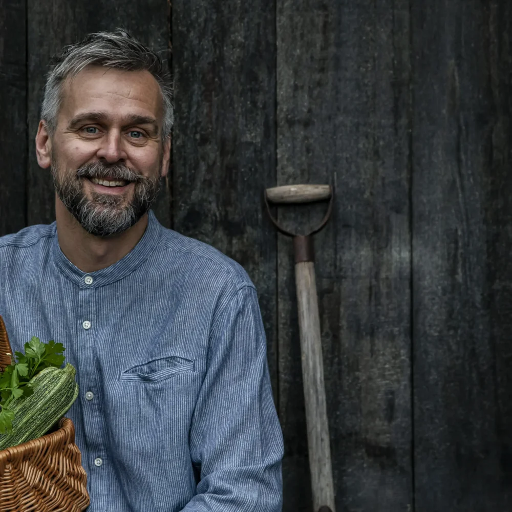 Bilden visar en person i en blå skjorta som håller en korg fylld med färska grönsaker, bland annat zucchini, mangold och morötter. I bakgrunden syns en mörk trävägg och en trädgårdsspade som lutar mot väggen.
