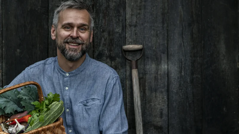 Bilden visar en person i en blå skjorta som håller en korg fylld med färska grönsaker, bland annat zucchini, mangold och morötter. I bakgrunden syns en mörk trävägg och en trädgårdsspade som lutar mot väggen.
