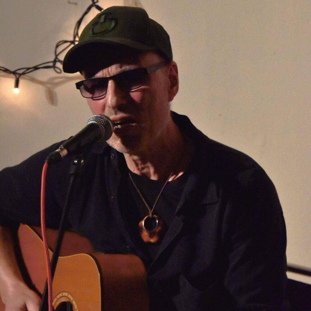 En man sitter och spelar gitarr. Han har solglasögon och keps