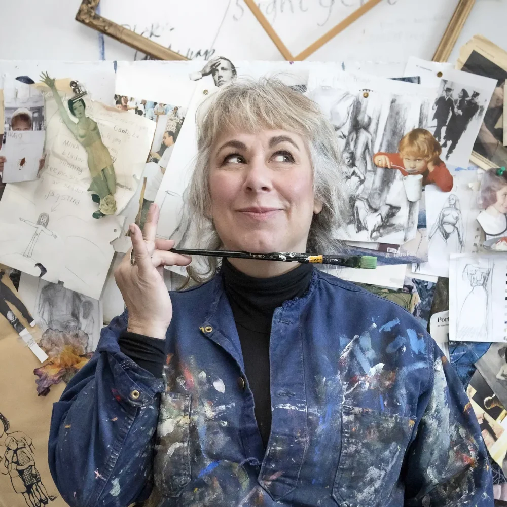 Stina Wollter står i blå målarrock och tittar snett uppåt höger hörn. under hakan håller hon en målarpensel.