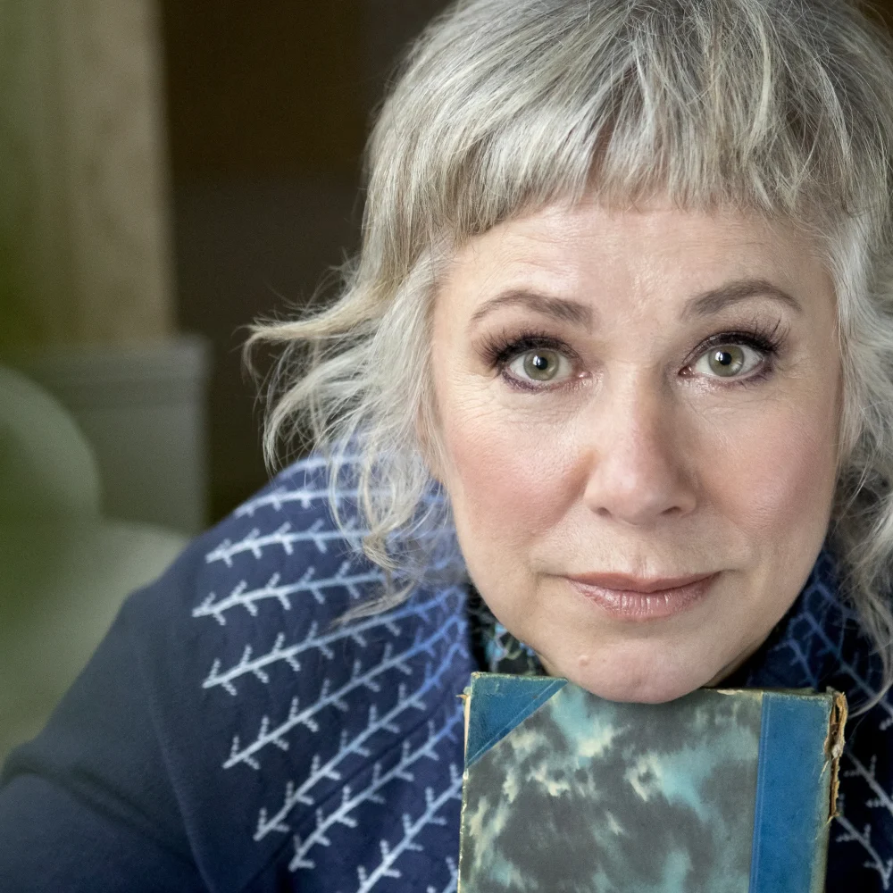 Stina Wollter sitter med en bok under hakan. Bär en blå stickad tröja och tittar in i kameran.