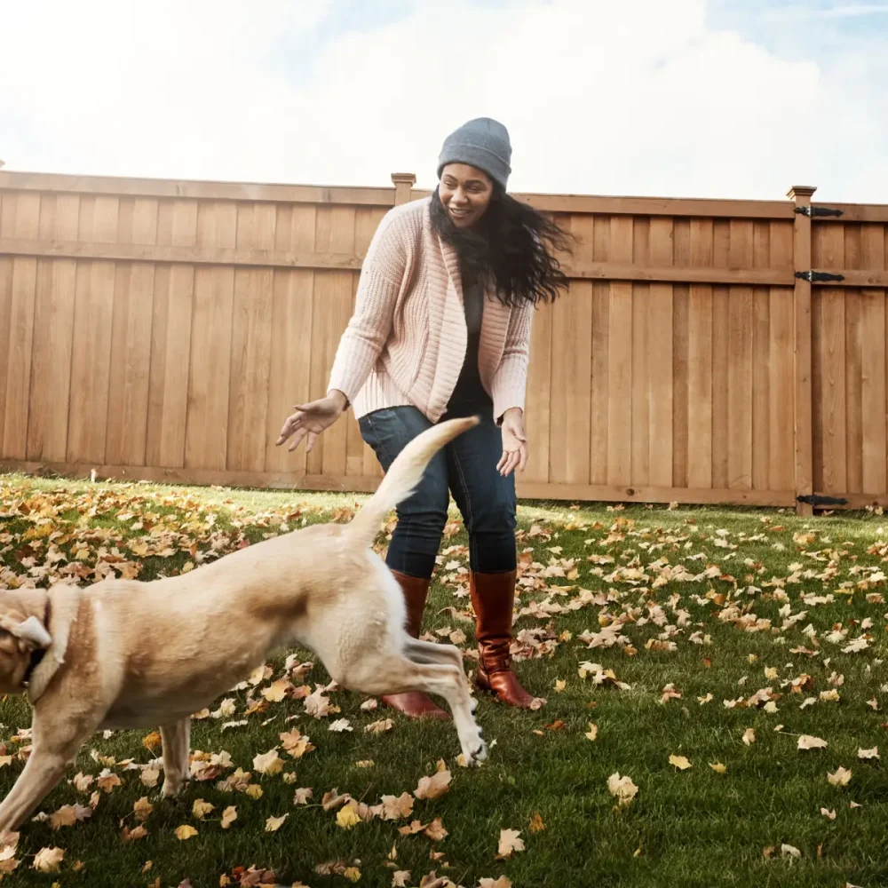 en kvinna i en mössa och ljuströja leker med en hund i trädgården